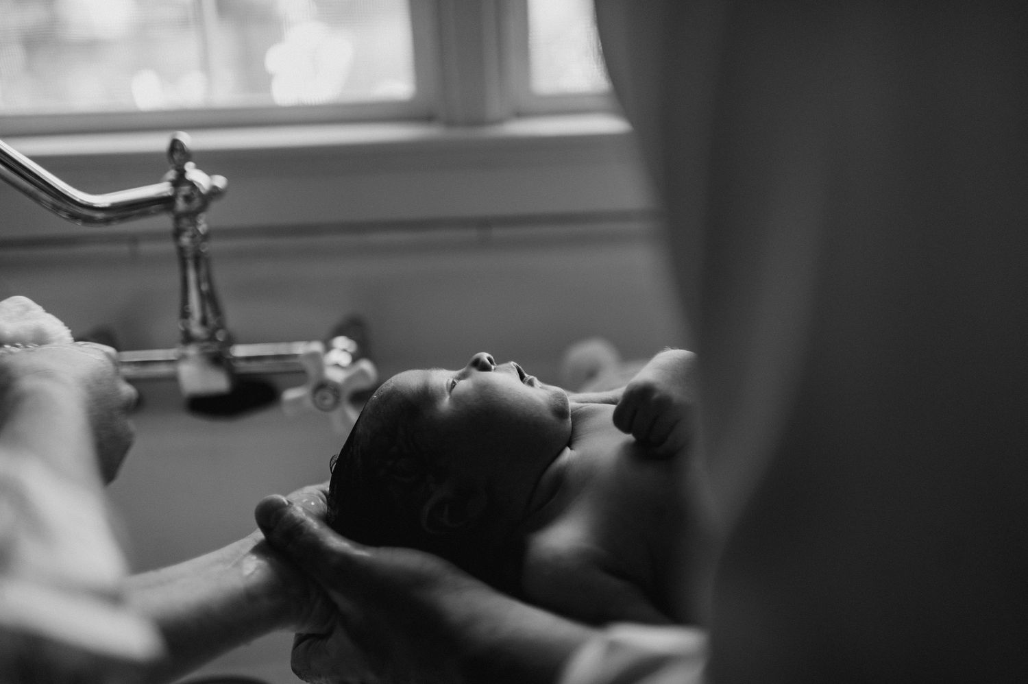 Black and white photo of a newborn baby being gently bathed in a sink by caring hands.