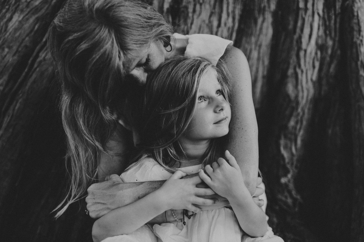 Black and white photo of a mother tenderly embracing her young daughter near a large tree trunk.