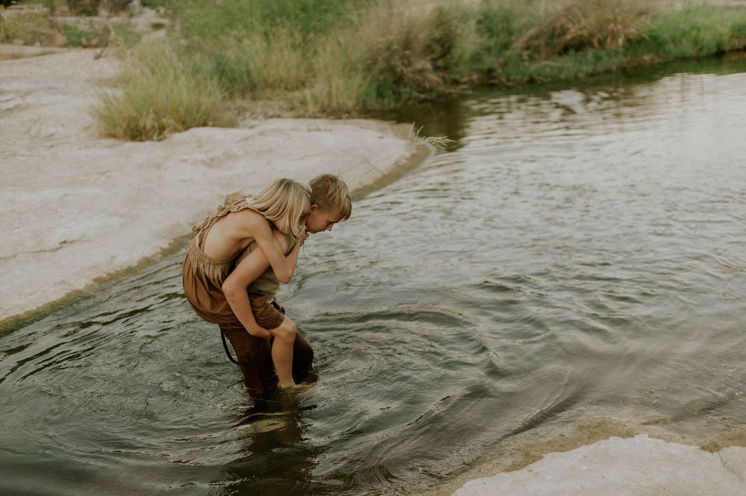 Two blonde children playing in a shallow river surrounded by nature.