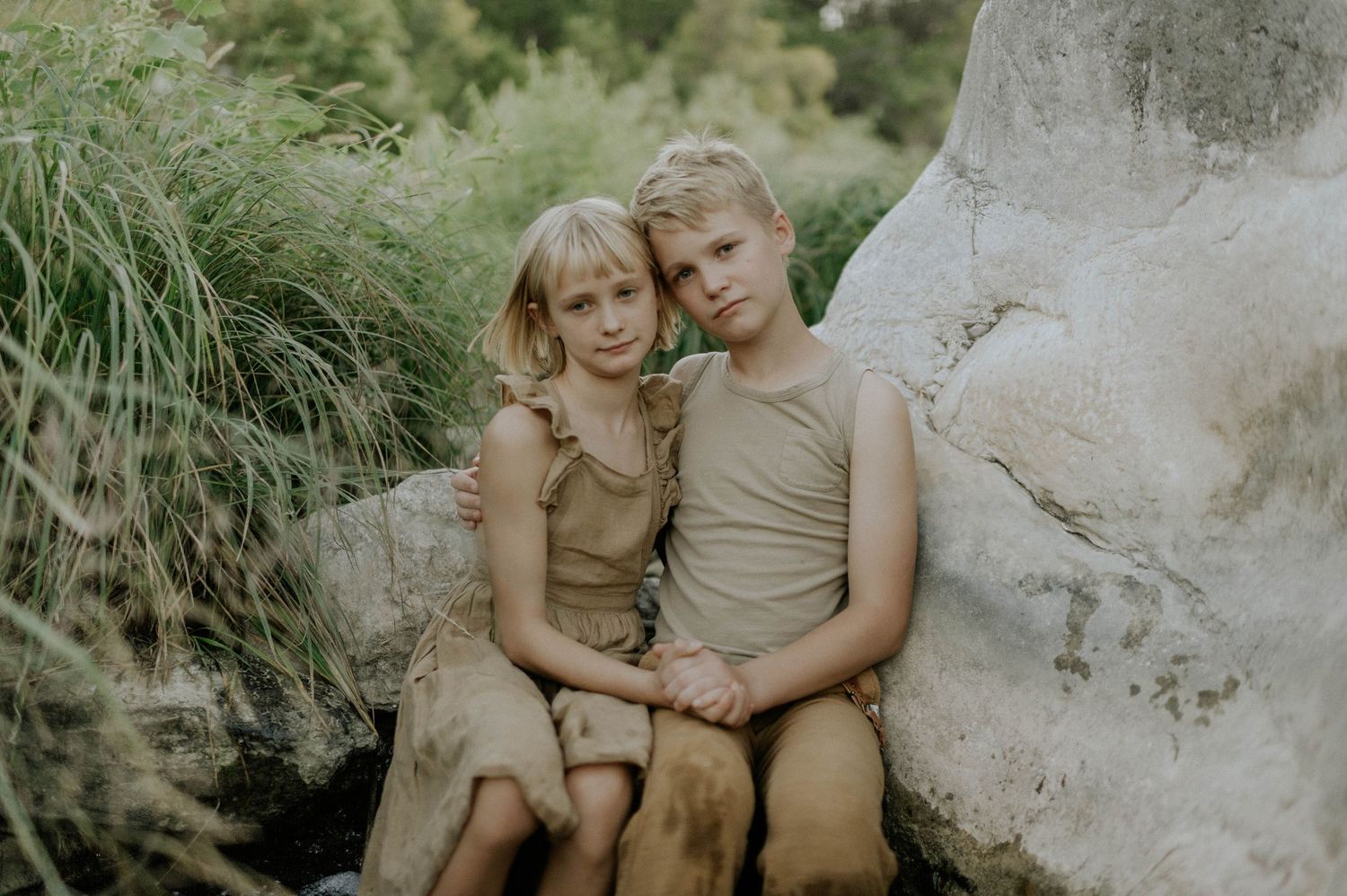 Two young blonde children in neutral outfits sit closely together near rocks and tall grass.