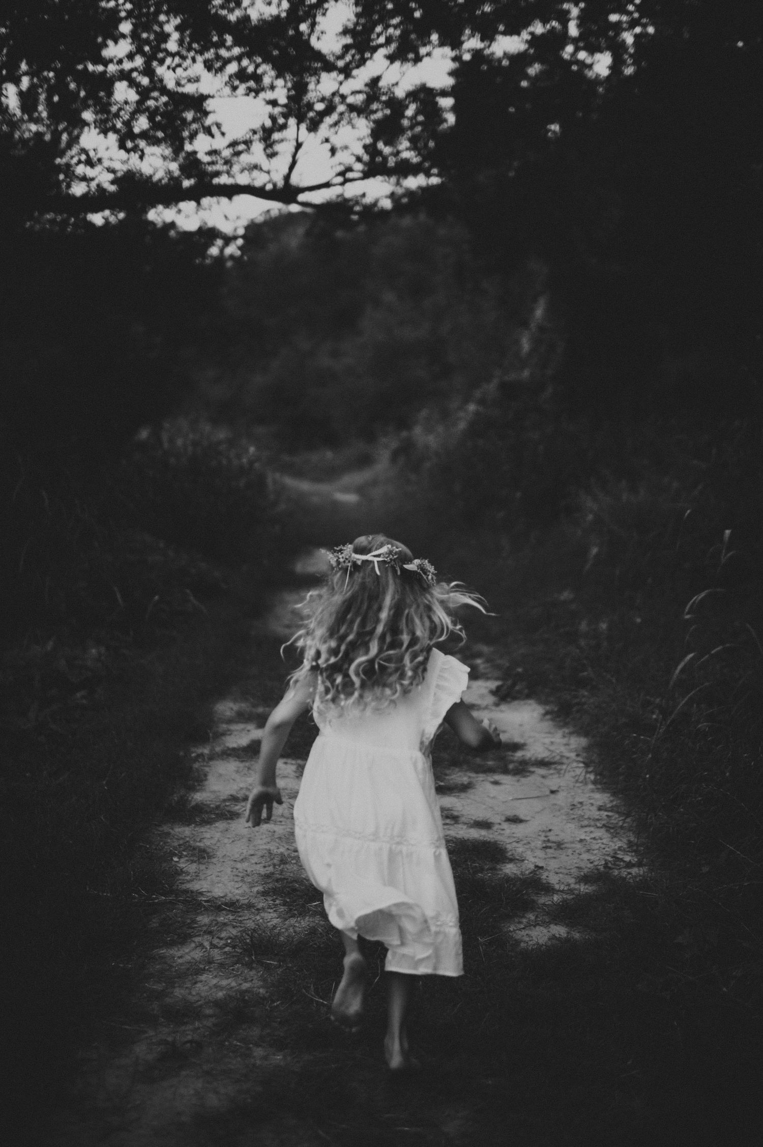 Young girl in white dress running barefoot down a dark forest path wearing a flower crown.