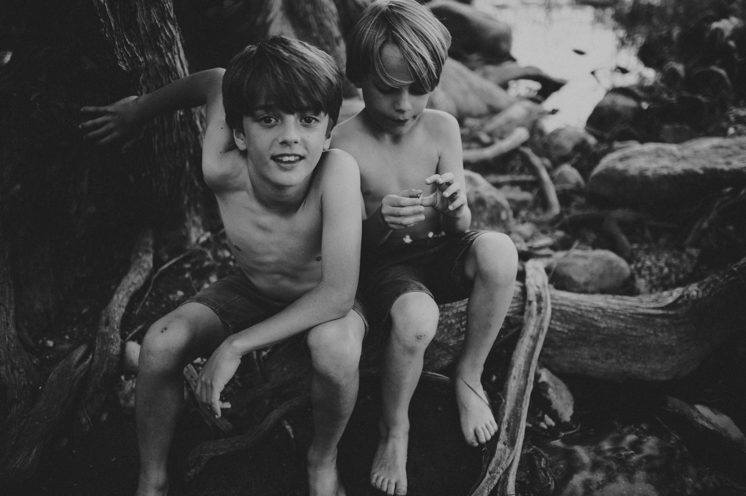 Two shirtless boys sitting on driftwood roots in a black and white outdoor nature photo.