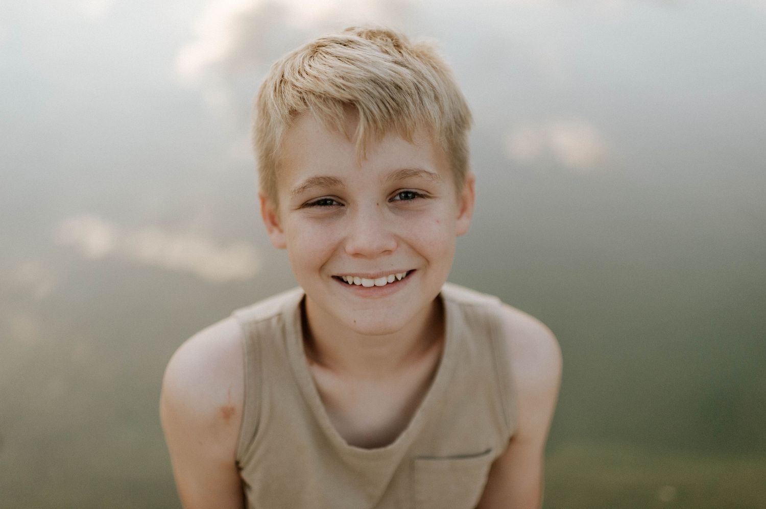 Smiling young blonde boy in a tan tank top posing near a misty lake.