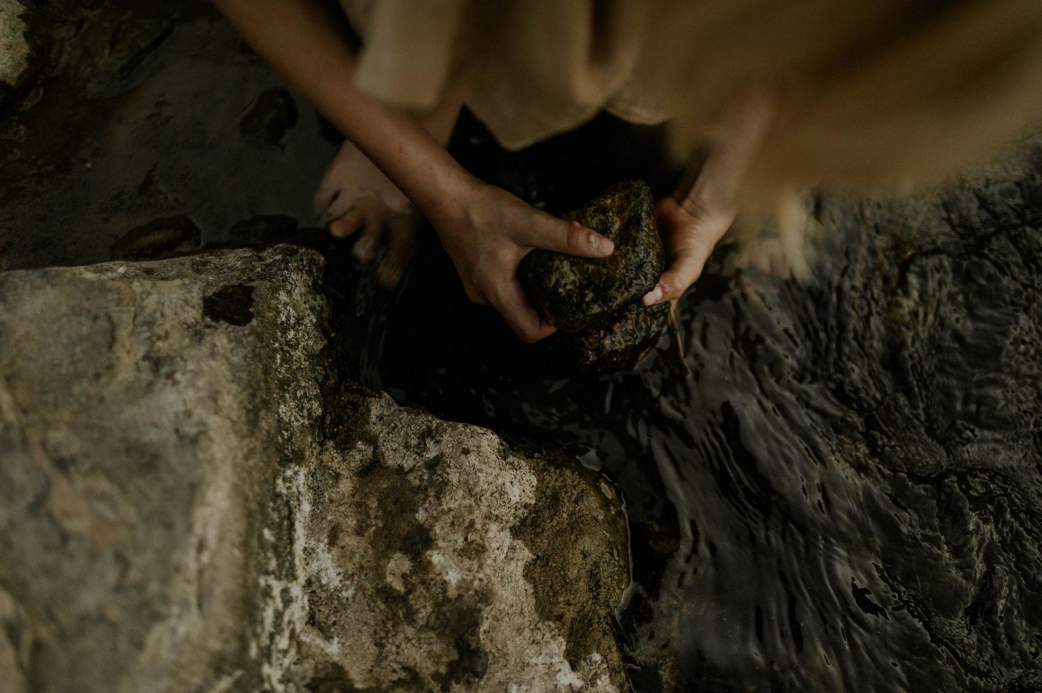 Child's hands holding a clump of dark wet mud beside a rocky stream in nature.