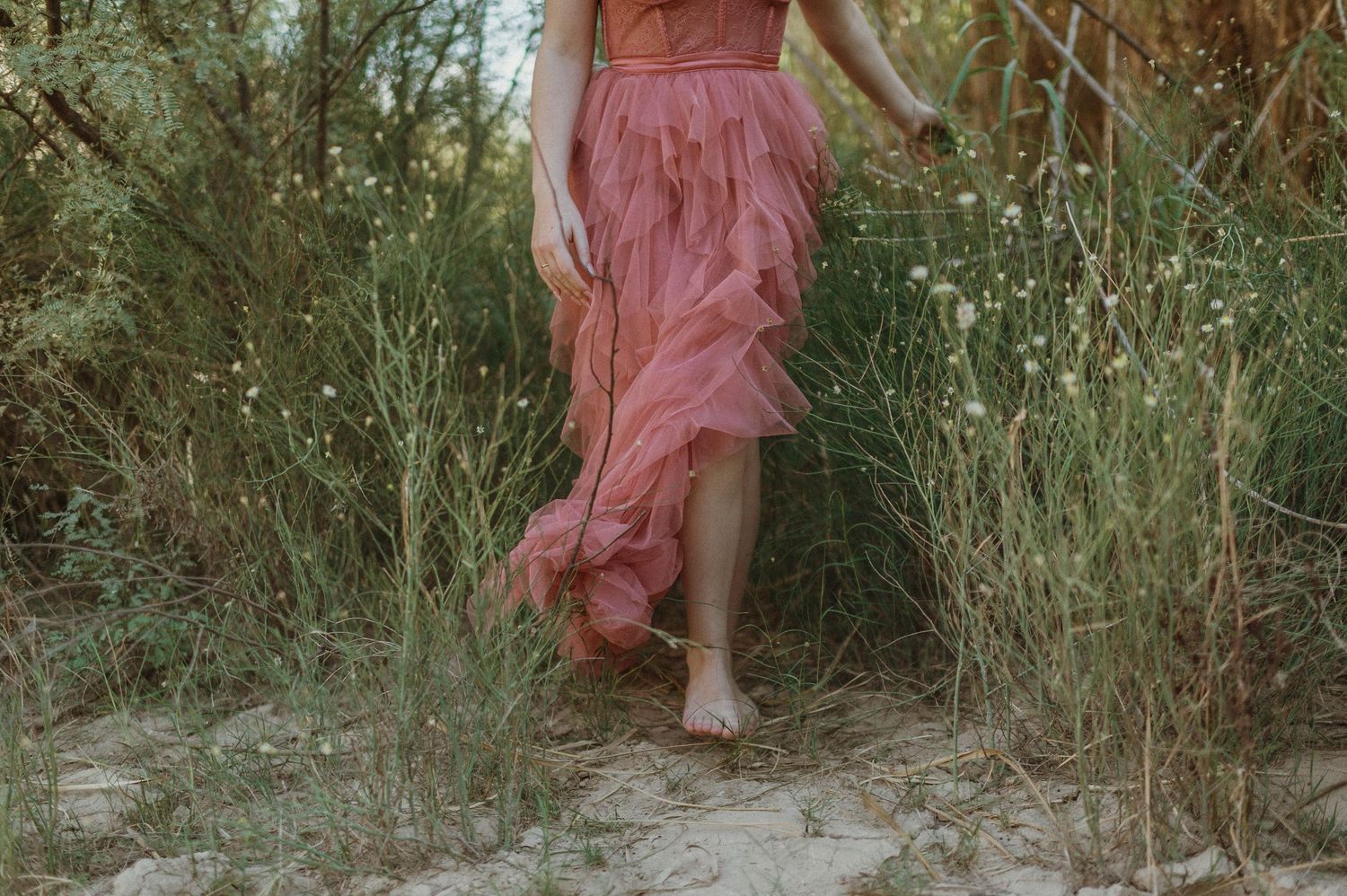 Barefoot woman in flowing pink ruffle maxi dress walks along sandy nature trail.