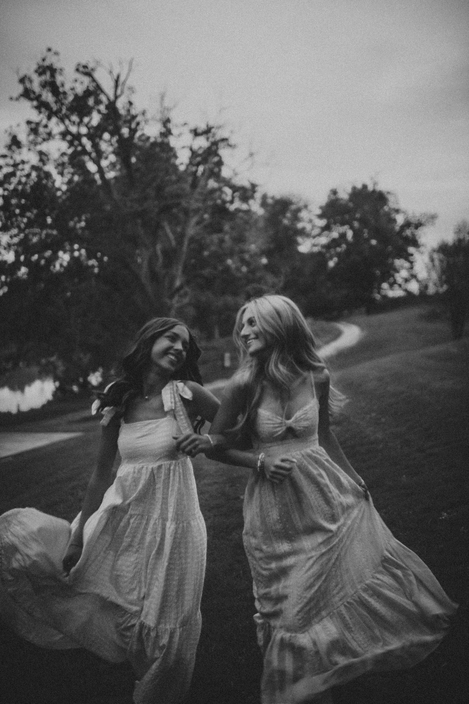 Two young women in flowing white dresses laughing and walking together outdoors in black and white.