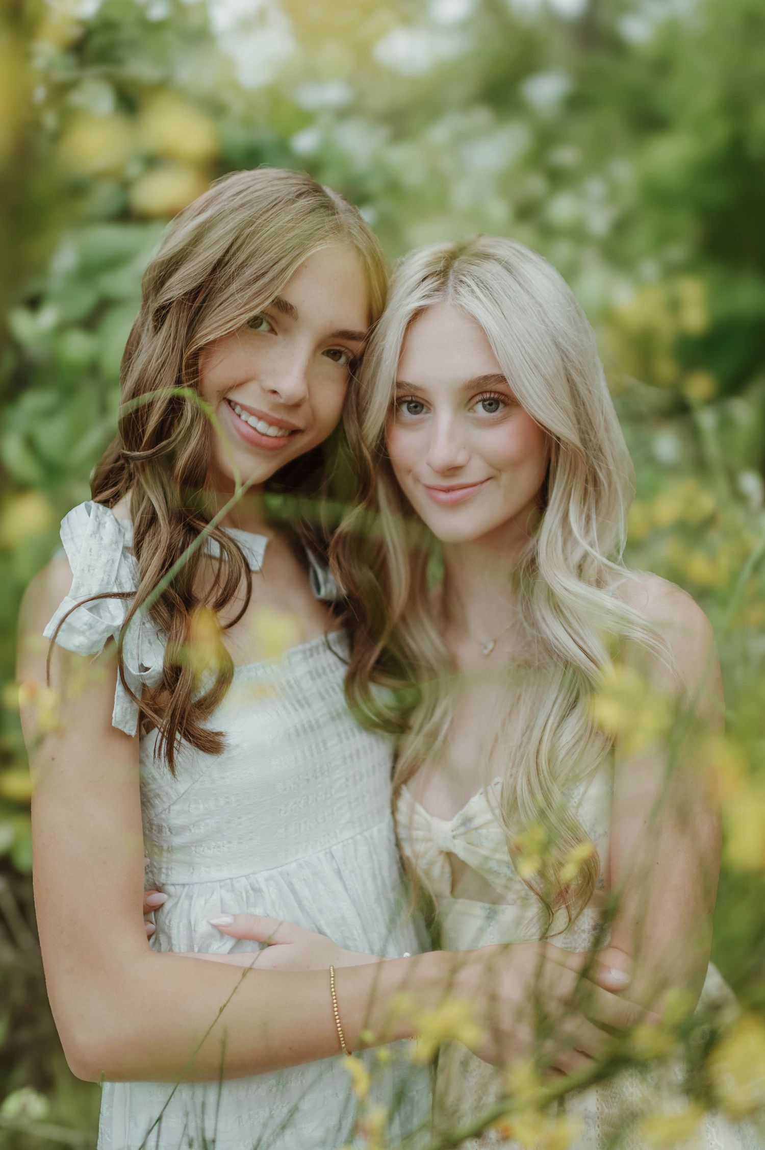 Two teenage girls in white dresses smiling together among yellow wildflowers in a bright outdoor portrait.