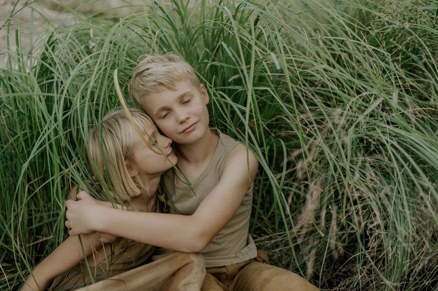 Two blonde children share a warm hug while sitting among tall green grass outdoors.