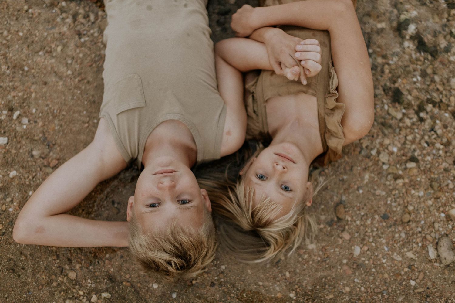 Two blonde children lying on sandy ground looking up at the camera.