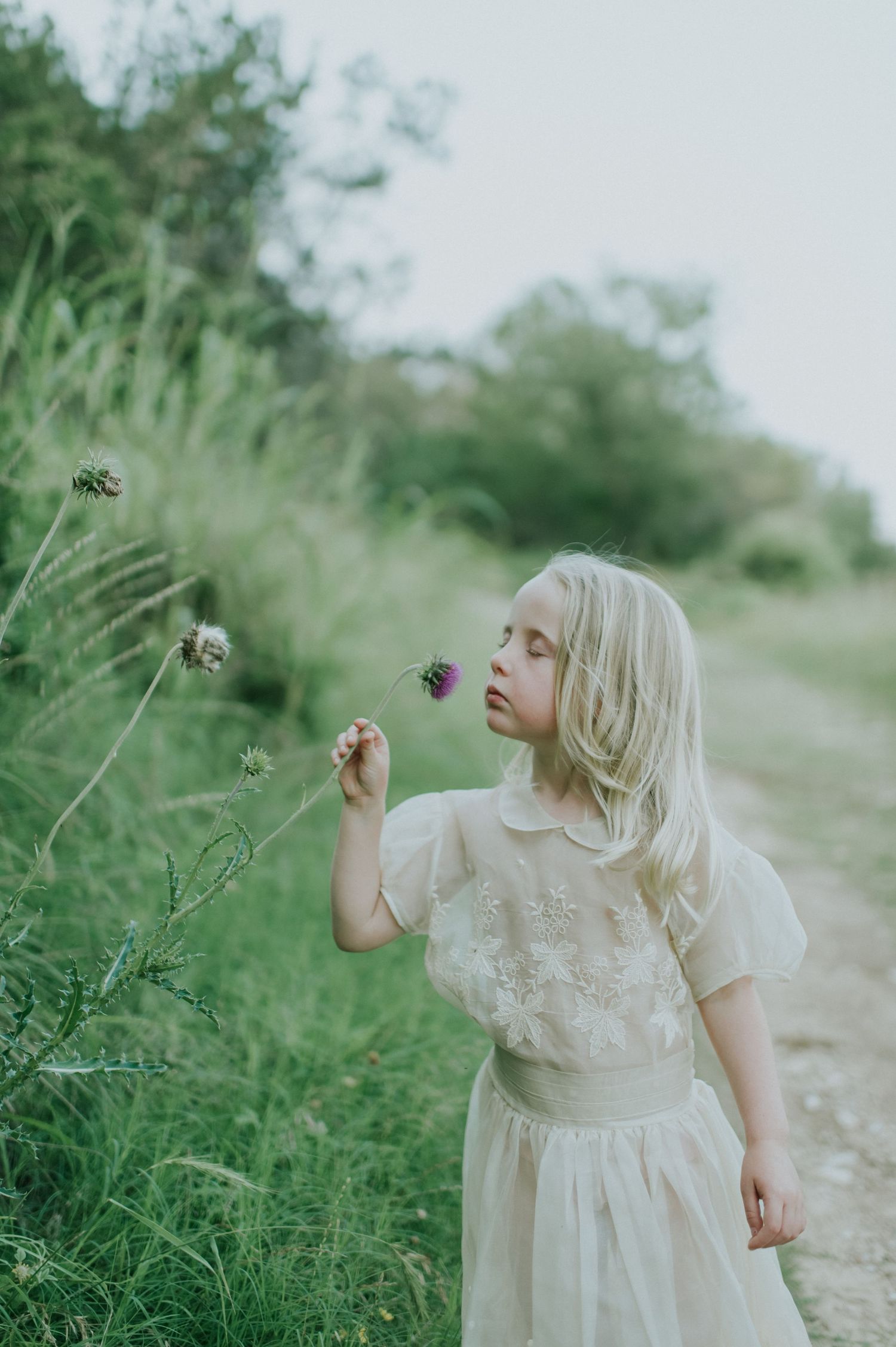 A young blonde girl in a white dress smells a wildflower on a lush green countryside path.
