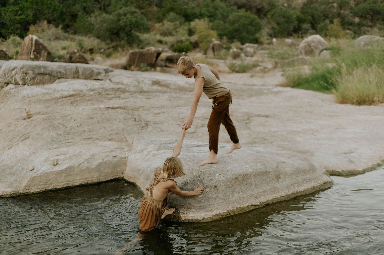 Boy helping young girl climb onto large boulder over a peaceful river in natural rocky landscape.