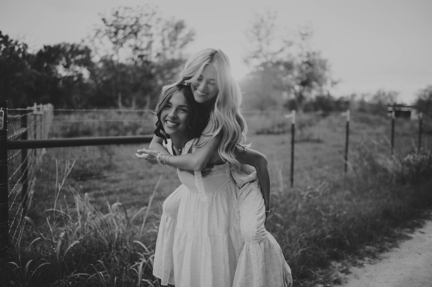 Two girls in white dresses share a joyful piggyback ride outdoors in a black and white photo.