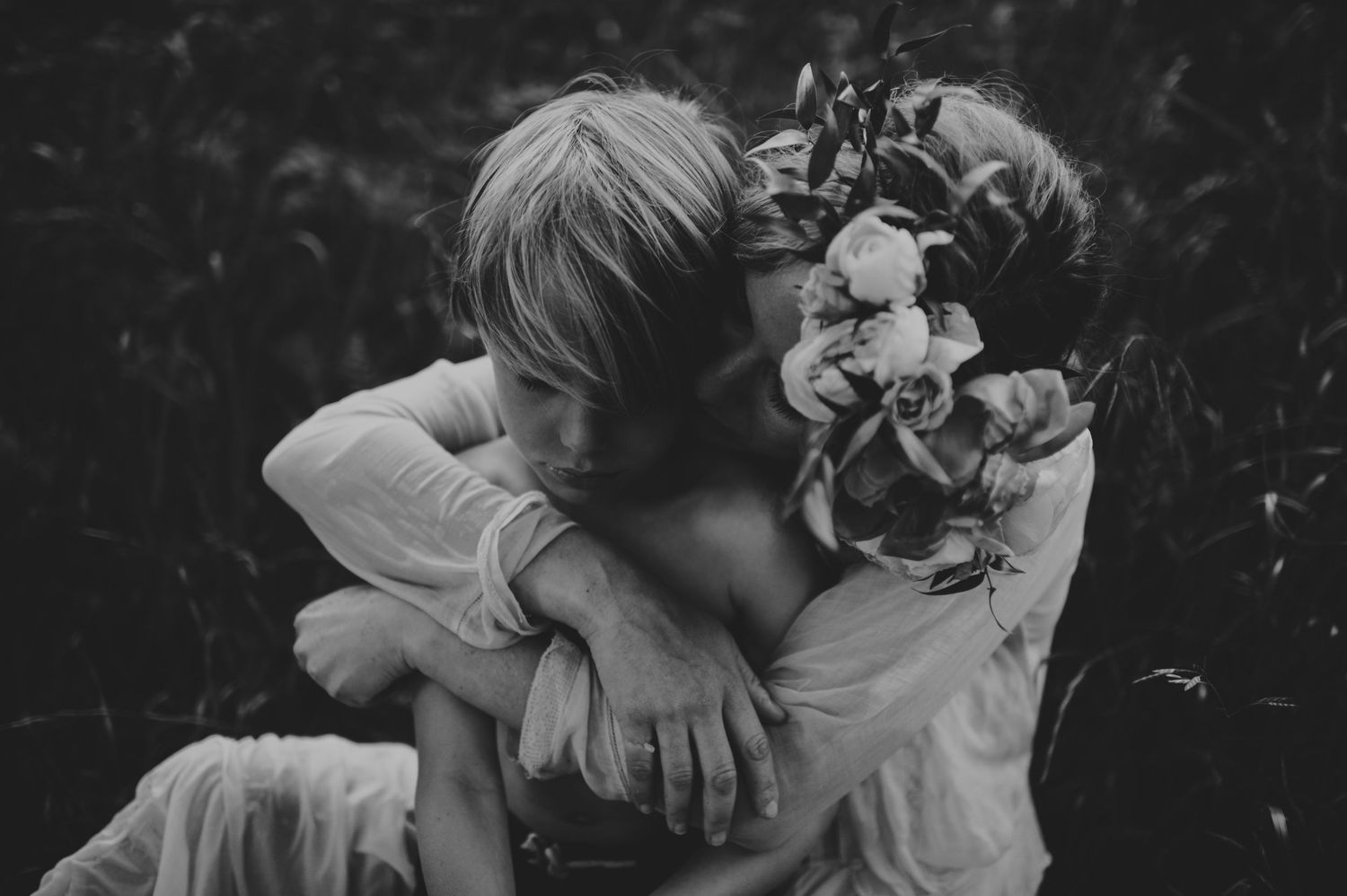 Two children sharing a tender hug outdoors, one wearing a floral crown, in black and white.