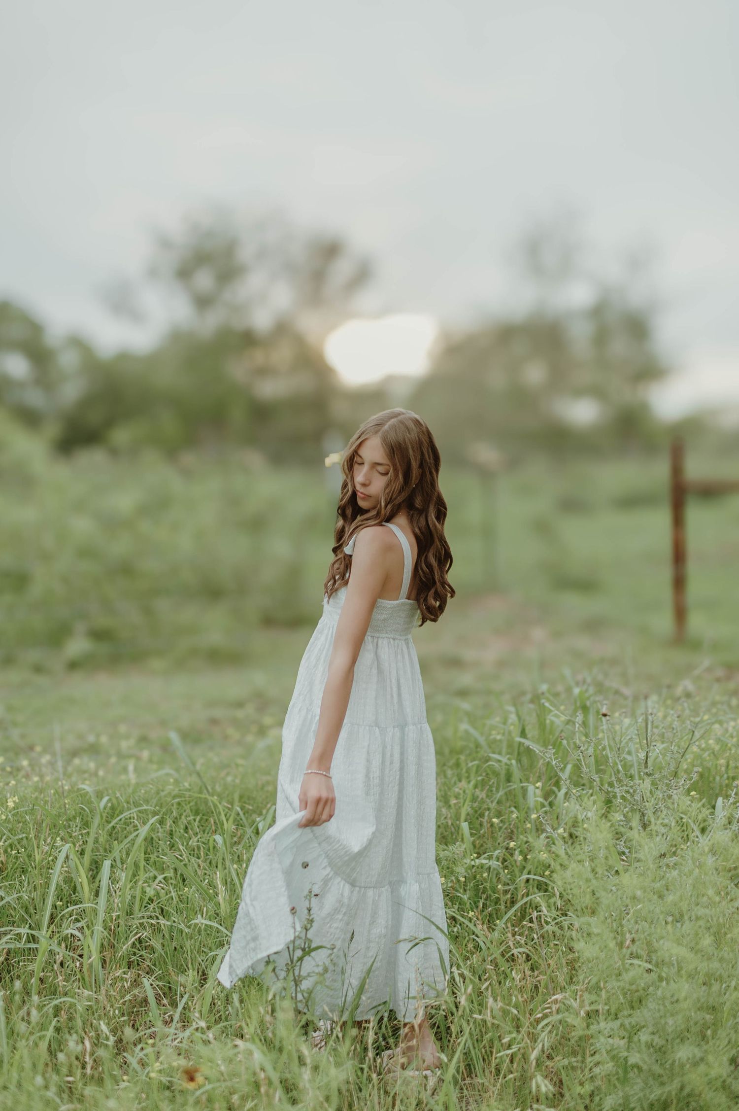 Young girl in a white maxi dress standing in tall grass during golden hour in a serene field.