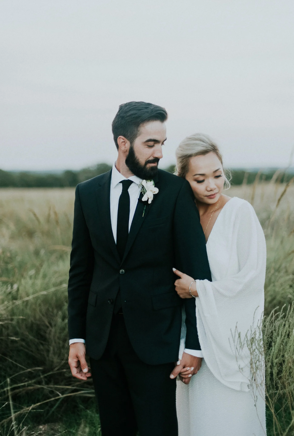 Bride and groom standing in a golden grass field, bride in flowing white dress, groom in black suit.