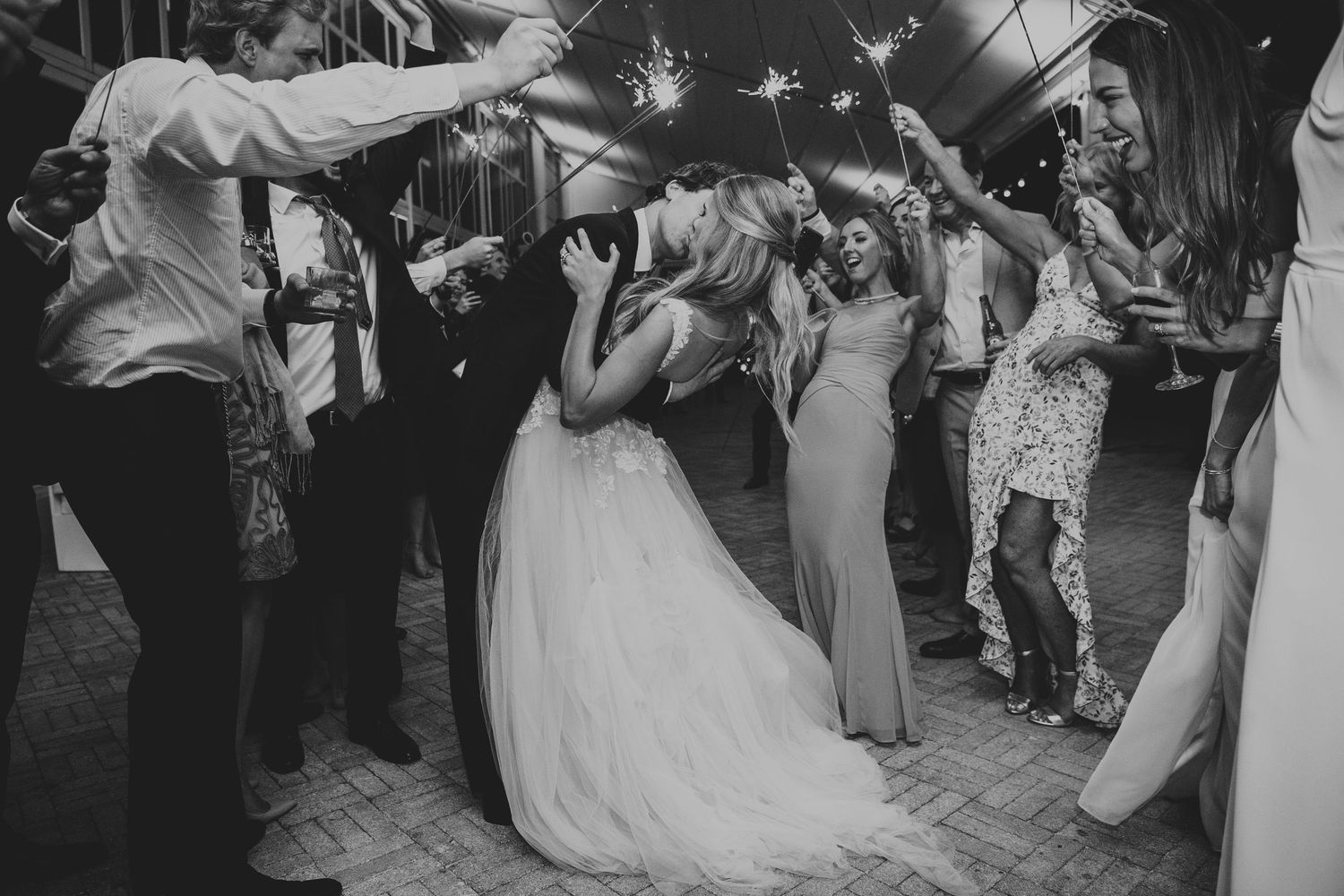 Black and white photo of bride and groom kissing surrounded by guests holding sparklers.