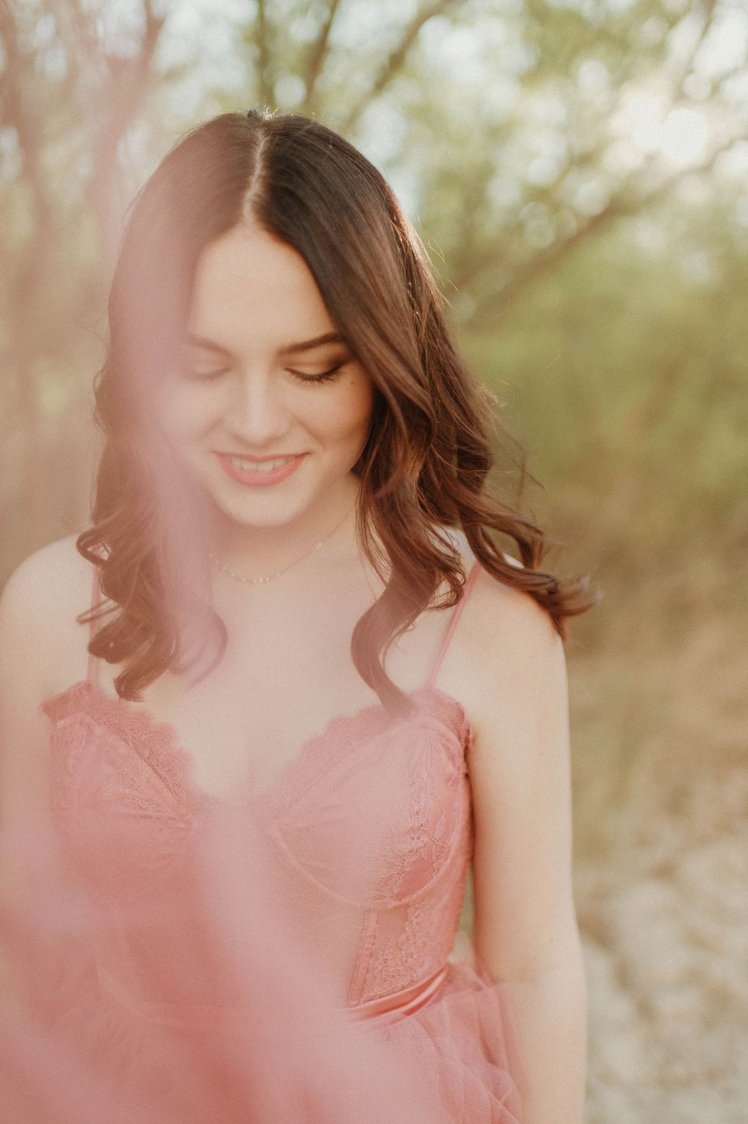 Smiling young woman in pink dress captured through soft dreamy tulle in natural setting.