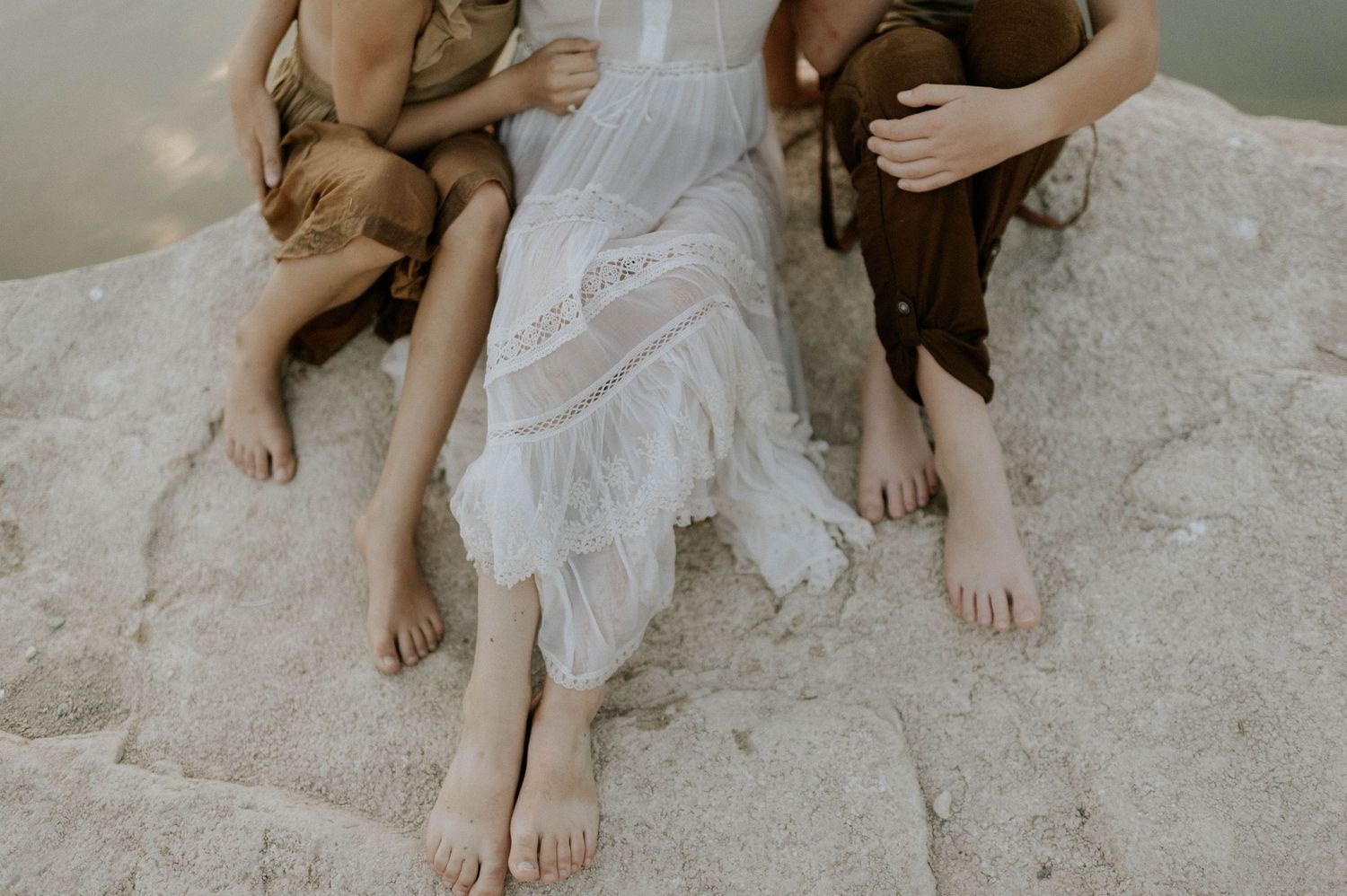A woman in a white dress sits barefoot on a rock between two children near a calm river.