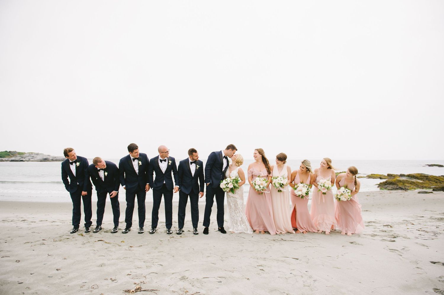 Wedding party walks along sandy beach, bridesmaids in blush gowns, groomsmen in navy tuxedos.