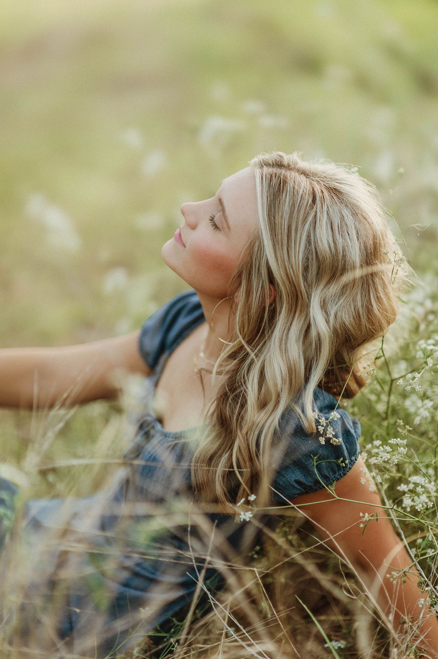 Blonde woman in blue dress sitting in wildflower field, head tilted back, enjoying golden hour sunlight.