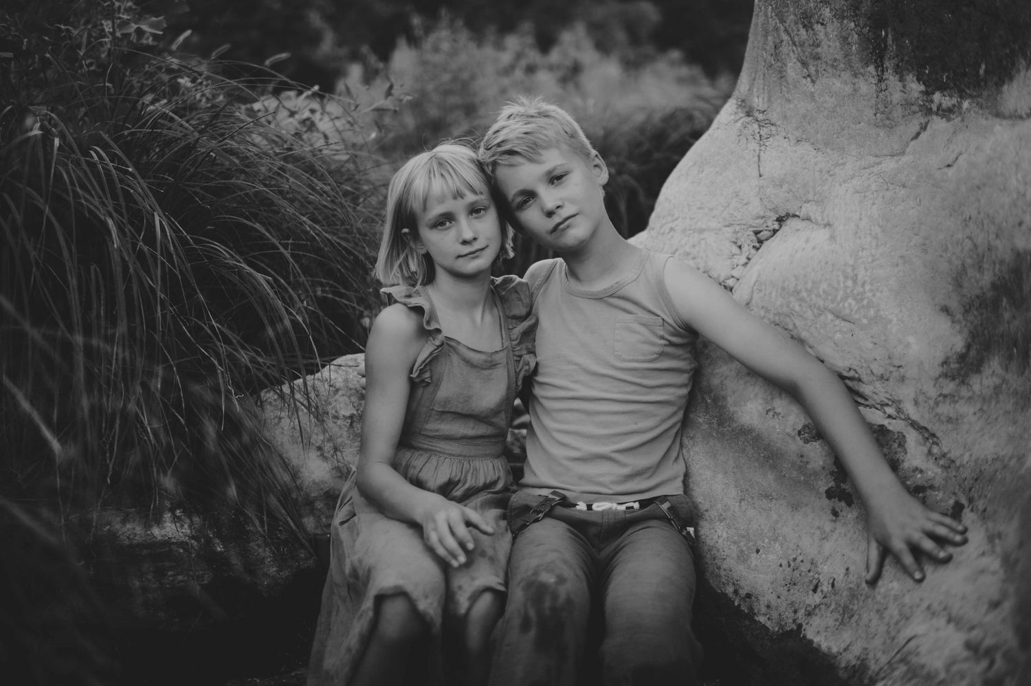 Two blonde children sit together leaning on a rock in a black and white nature photo.