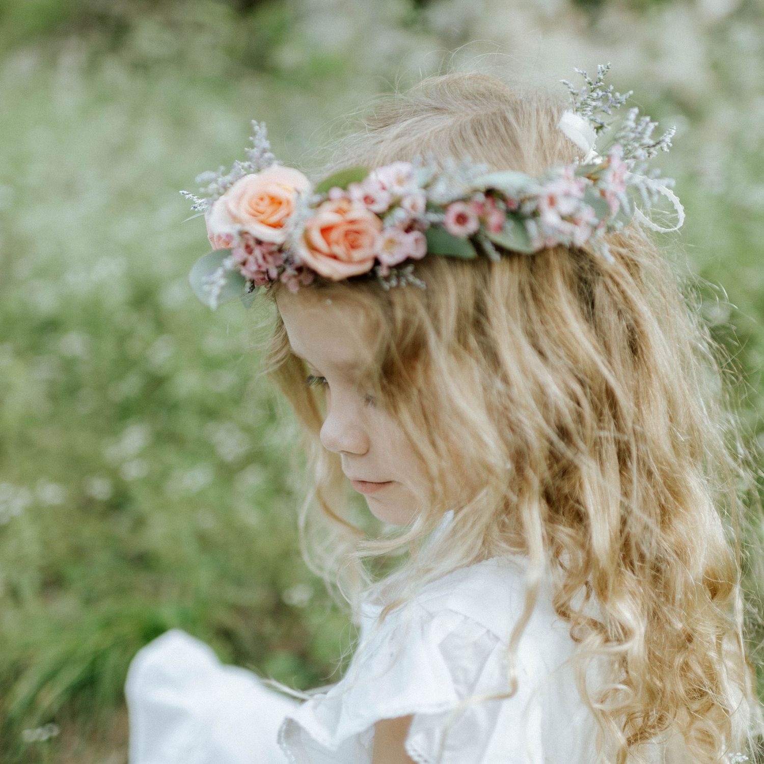 Young girl with long blonde hair wearing a floral crown of pink and peach roses in a garden.
