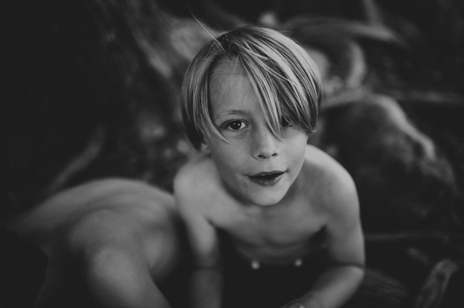 Black and white portrait of a shirtless young boy with blonde hair looking at the camera outdoors.