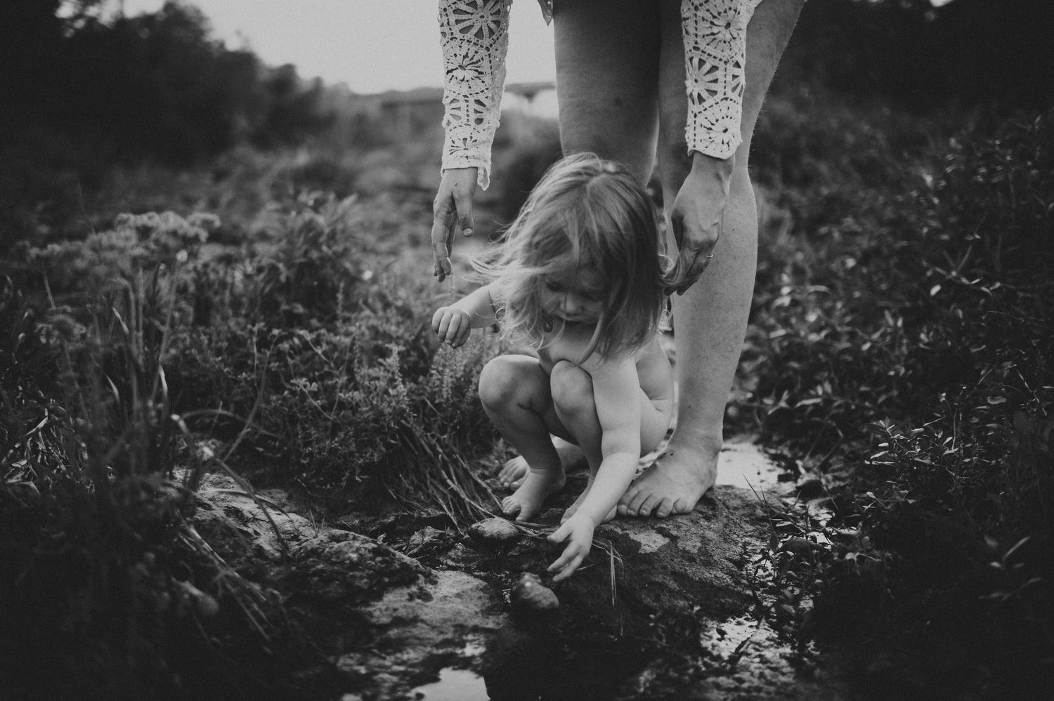 Black and white photo of a child exploring a muddy stream with an adult guiding them outdoors.