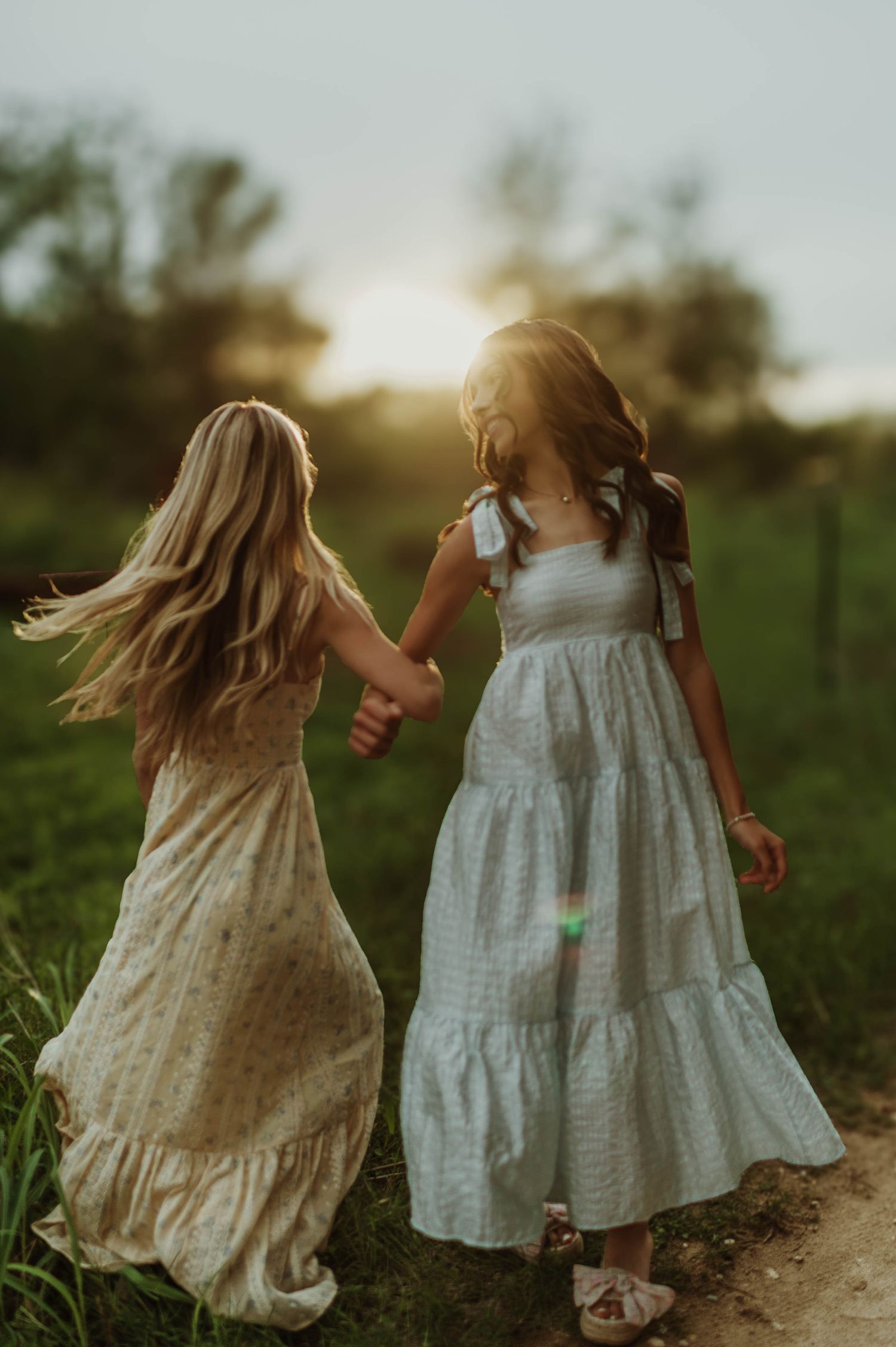 Two girls in flowy dresses holding hands walking toward a glowing sunset on a scenic outdoor path.