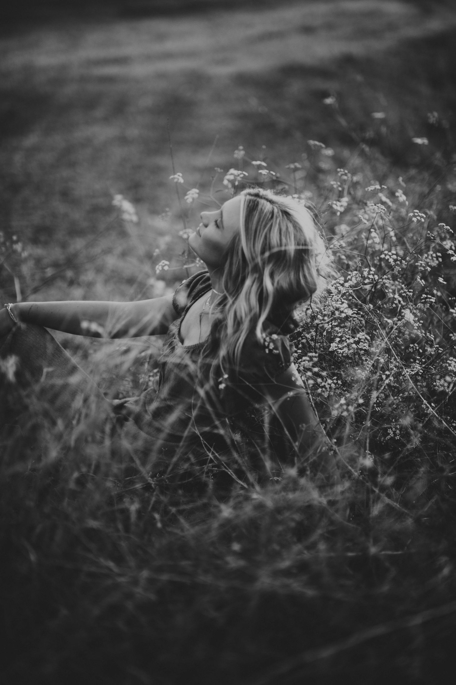 Black and white photo of a woman with long hair sitting in a field of wildflowers, gazing upward.