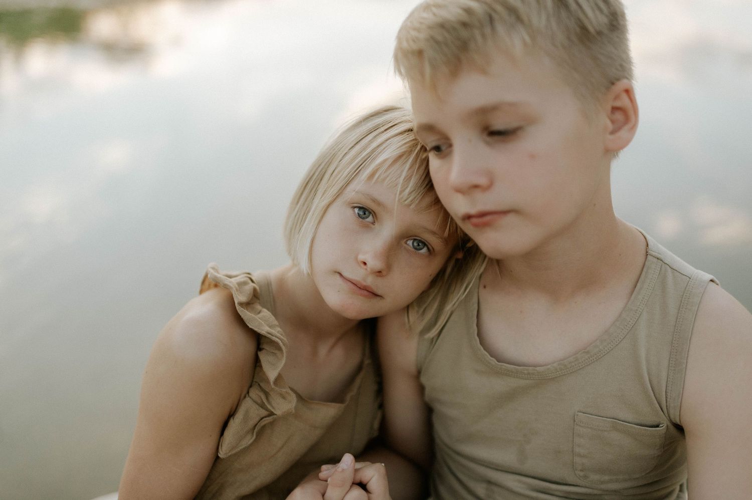 Young blonde girl resting her head on her older brothers shoulder near a lake.