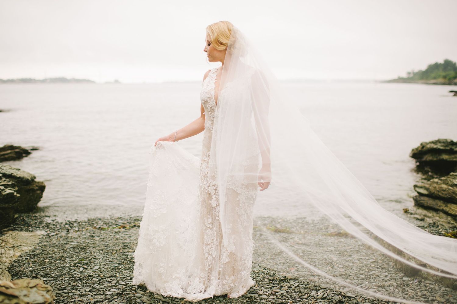 Bride in lace wedding gown and veil poses gracefully by the ocean shore.