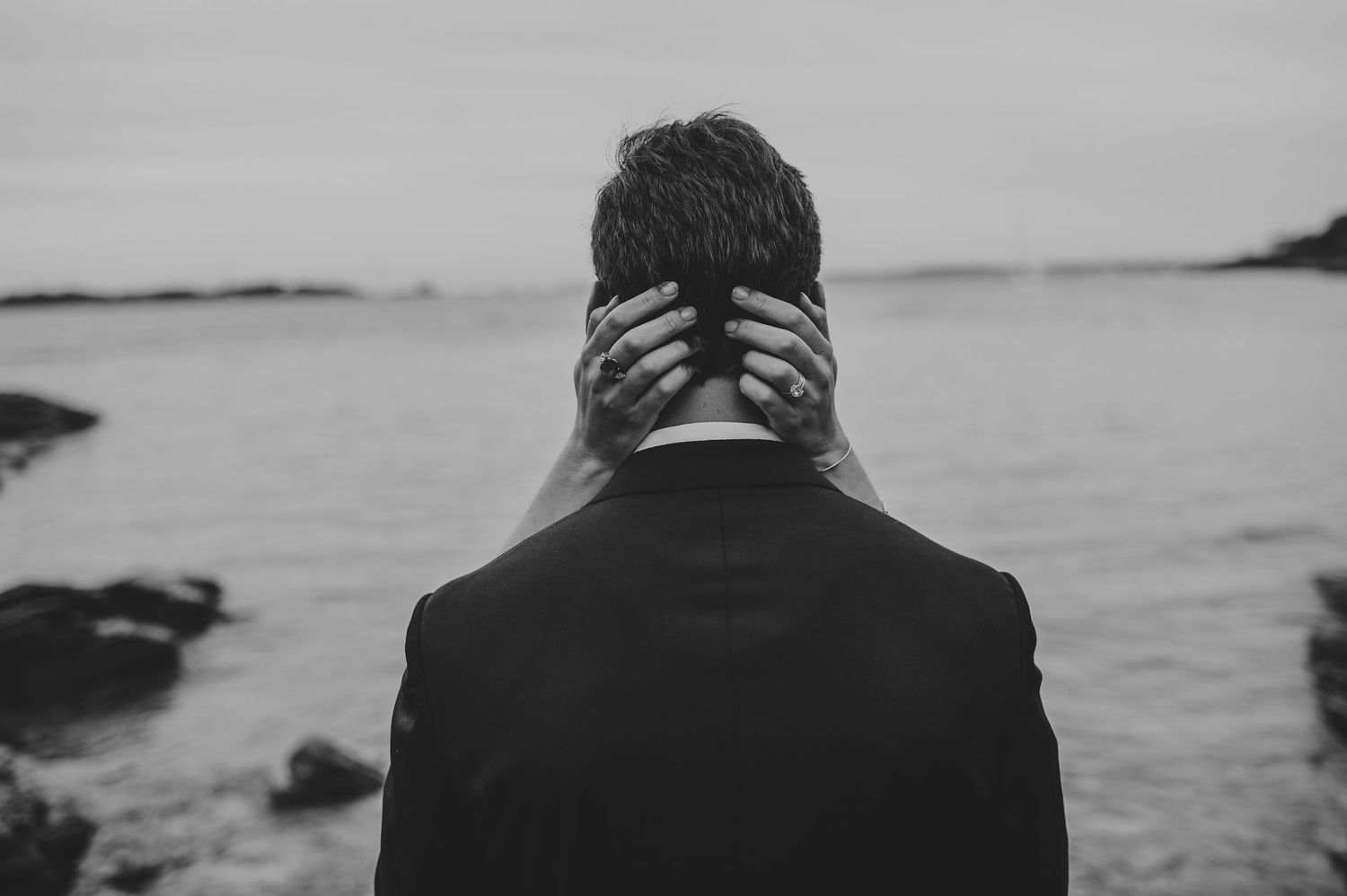 Black and white photo of groom standing at rocky shoreline with hands behind his head.