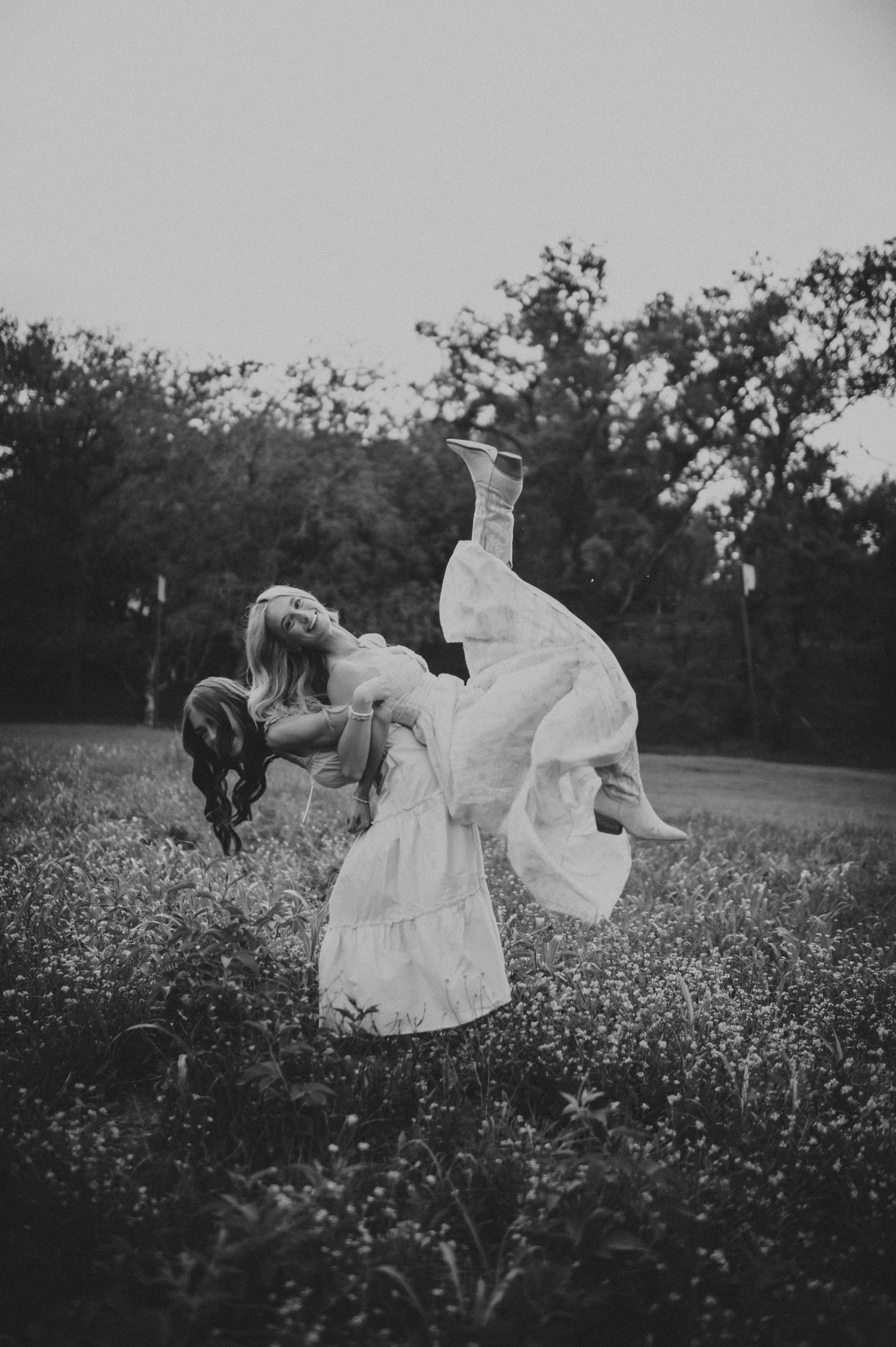 Black and white photo of a groom lifting his bride in a flower field with trees in the background.
