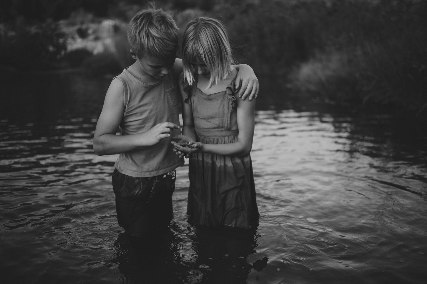 Boy and girl standing together in a river, boy holding something small while girl embraces him.