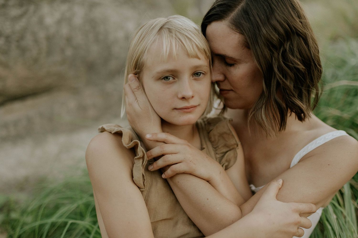 A brunette woman gently holds a young blonde girl close in a loving outdoor embrace.