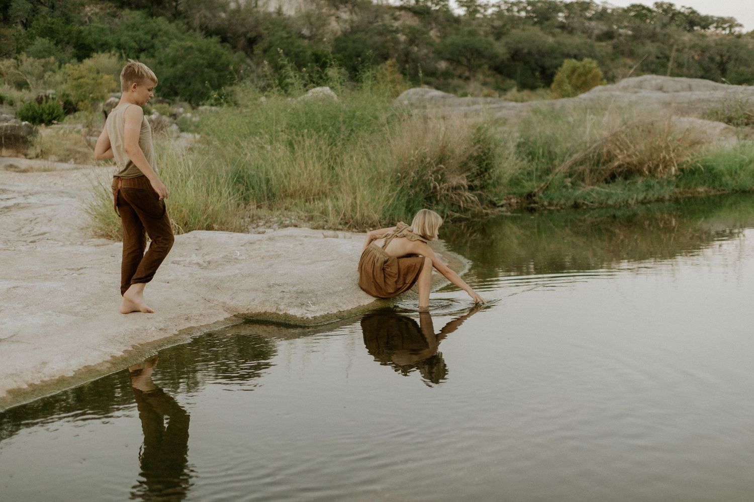 Two children playing by rocky riverbank, one crouching to touch the calm clear water.