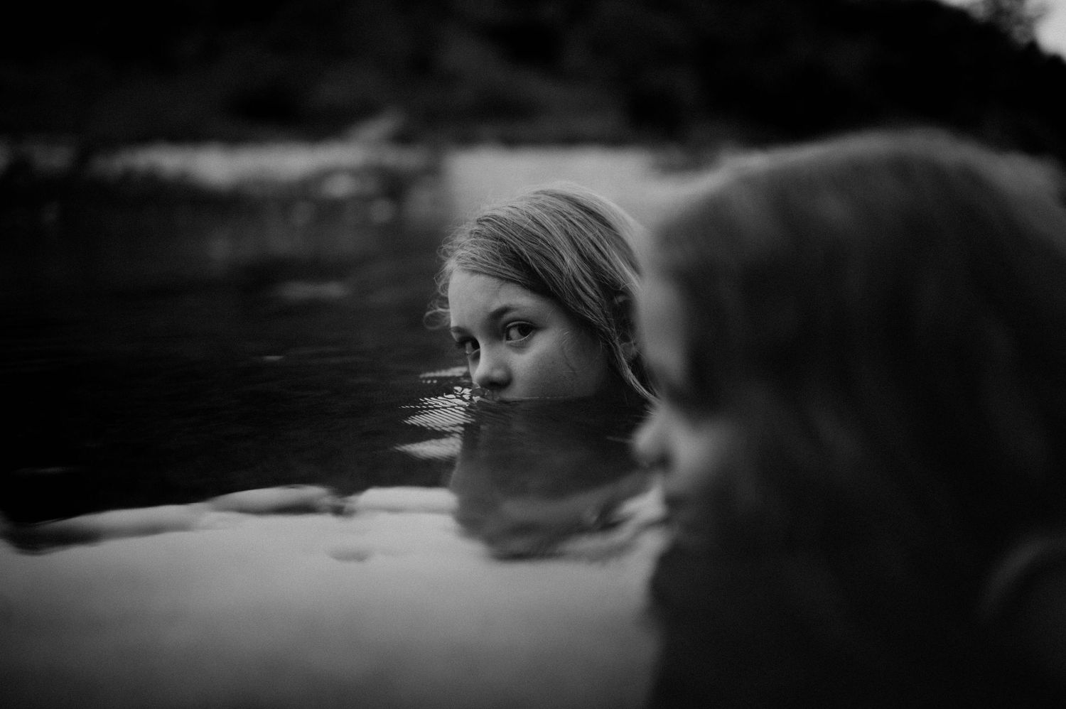 Black and white photo of a girl submerged in water up to her eyes, gazing at another person in the foreground.