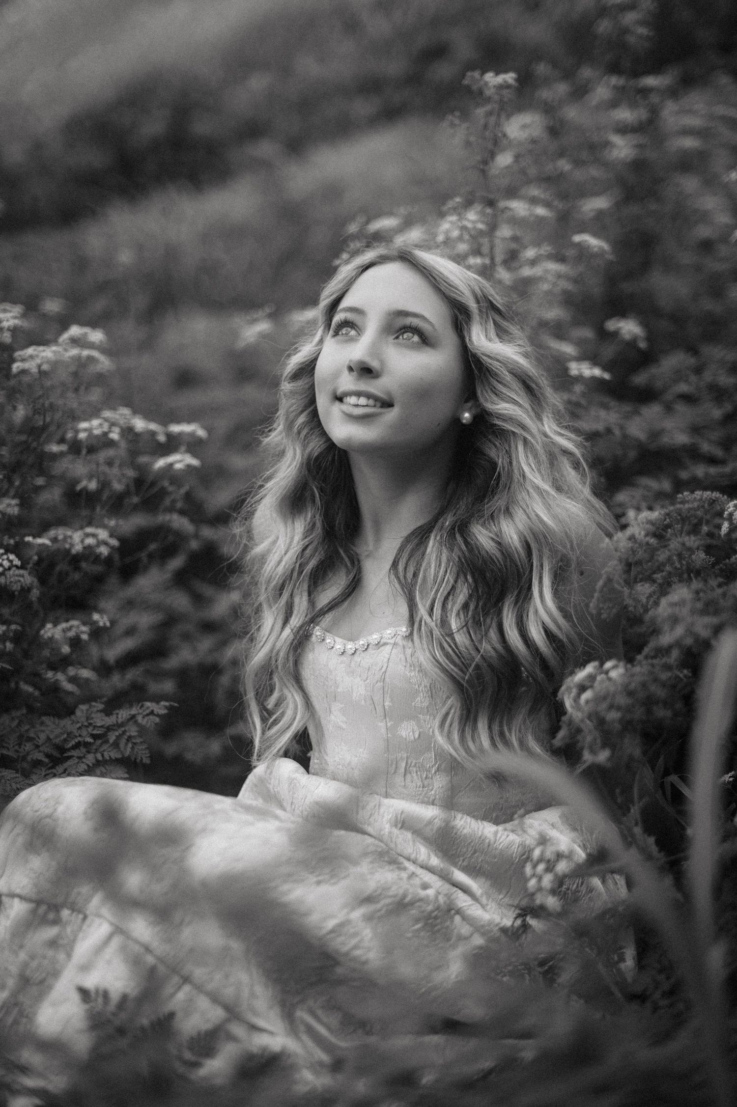 Black and white photo of a young blonde woman in a vintage dress sitting among wildflowers, gazing upward.