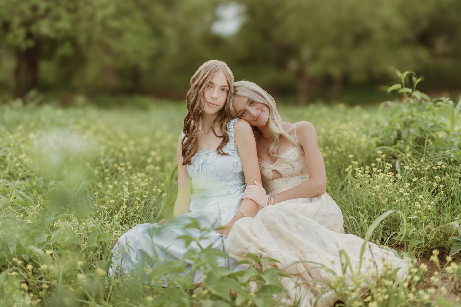 Two young women in pastel dresses sitting together in a lush green wildflower meadow.