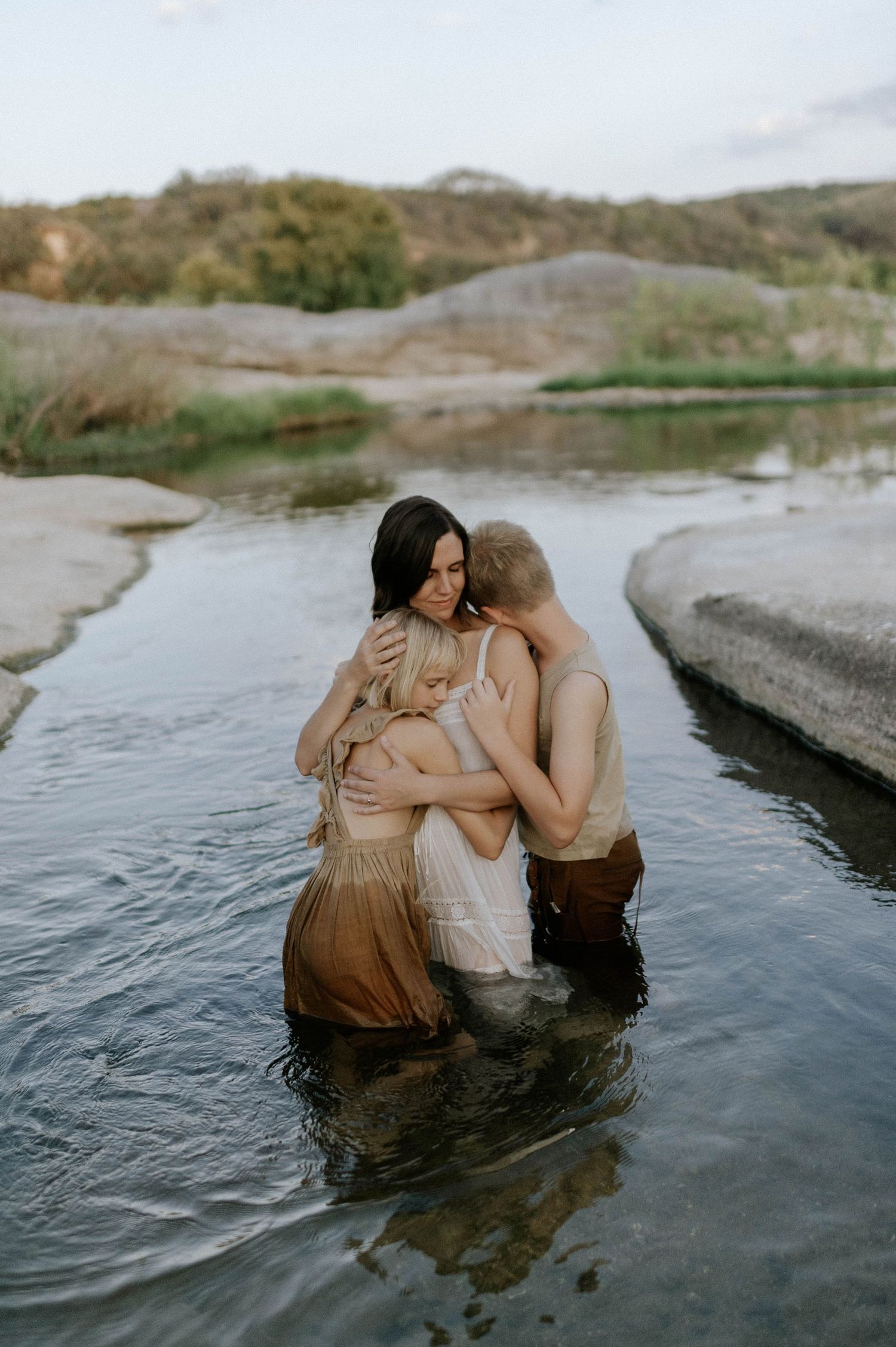 Mother embracing two children in a river with rocky landscape in soft natural light.