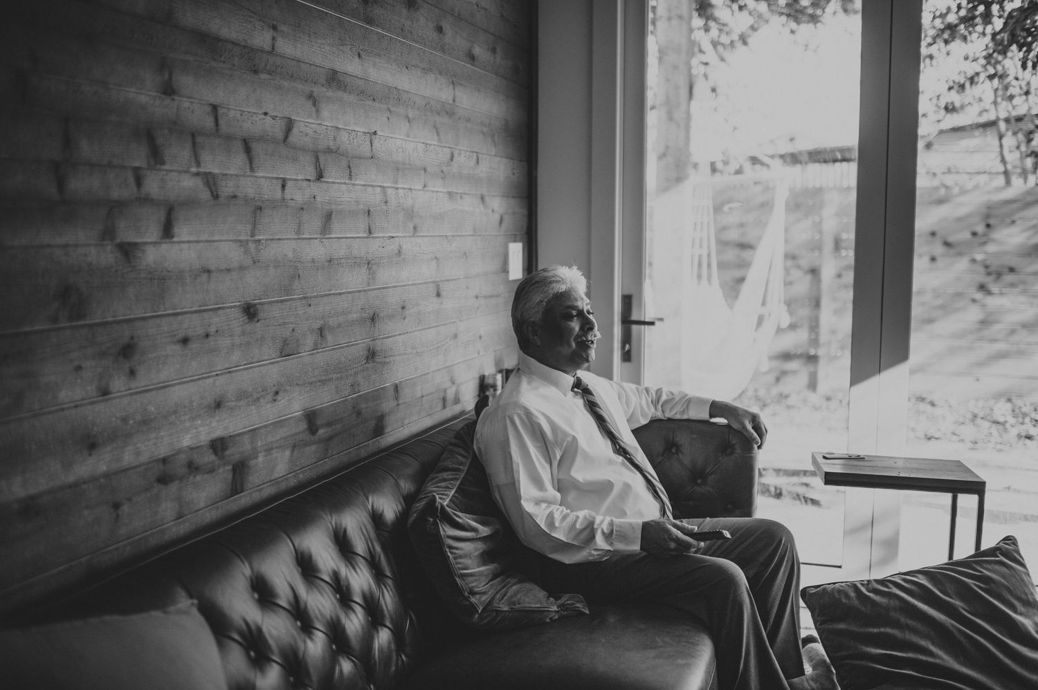 Elderly man in dress shirt sits contemplatively on leather sofa beside large window.