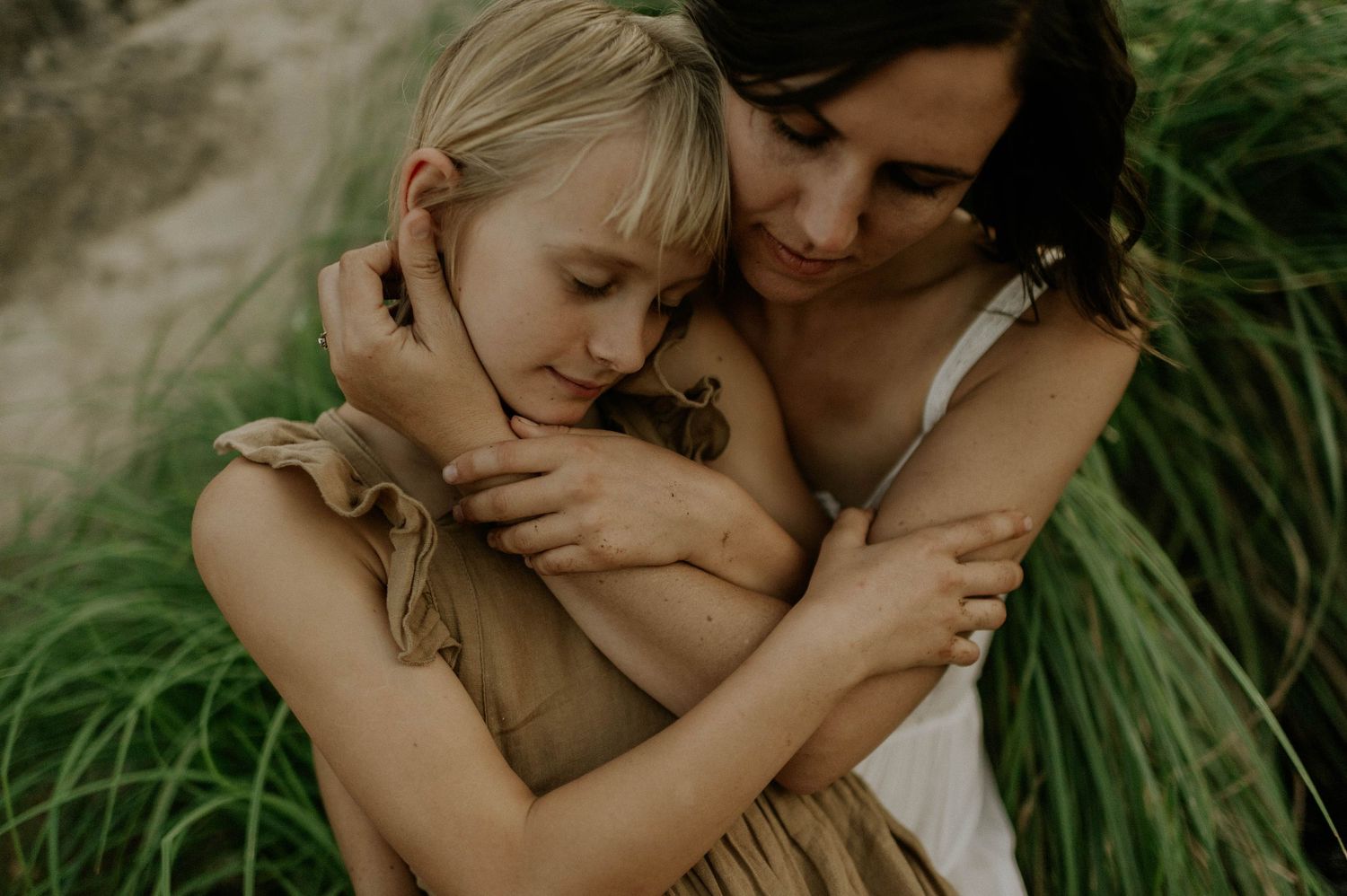 A dark-haired mother tenderly embraces her blonde daughter outdoors among green grass.