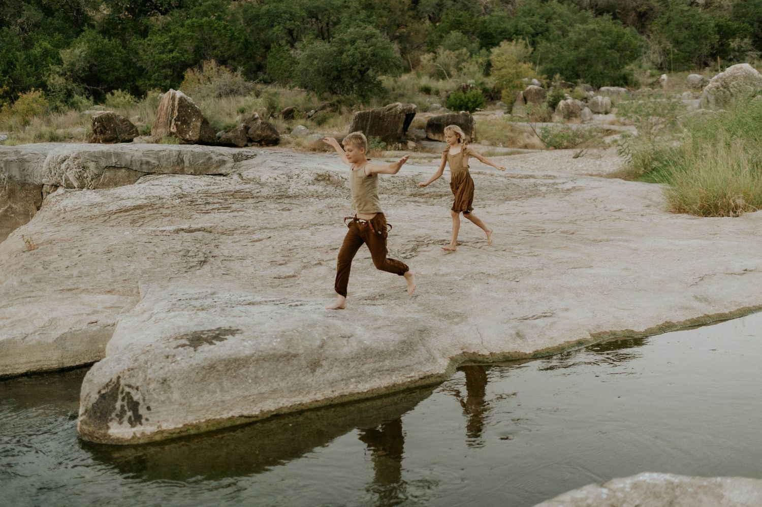 Two children running and jumping on large rocks beside a serene river in natural landscape.