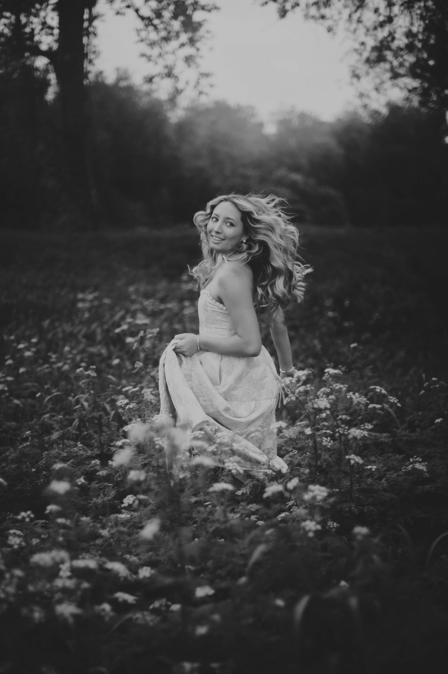 Black and white photo of a woman in a white dress sitting in a wildflower field with hair flowing in the wind.