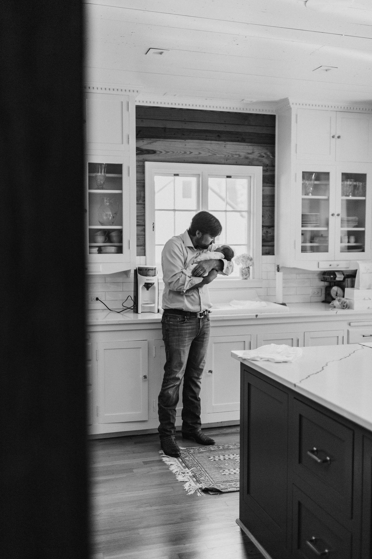 Black and white photo of a father tenderly holding his newborn baby in a bright white kitchen.