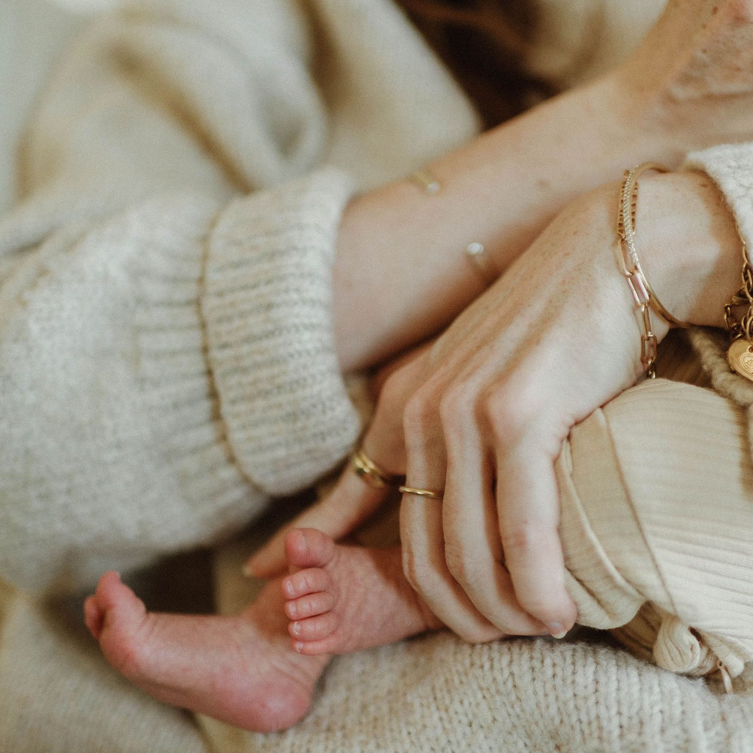 A mother in a cream knit sweater with gold bracelets gently holds her newborn baby's tiny feet.