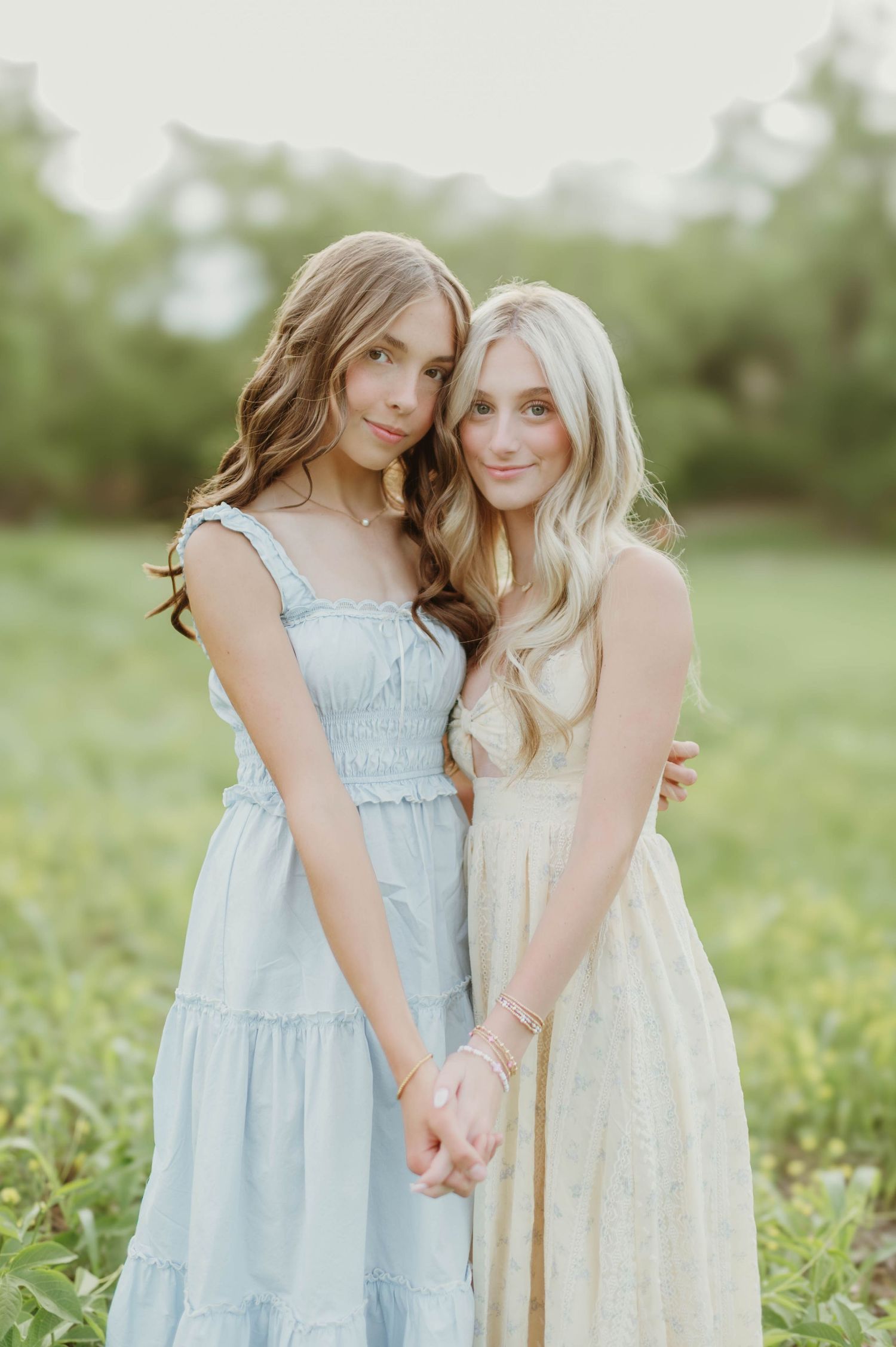 Two young women in blue and cream dresses holding hands in a green meadow during golden hour.