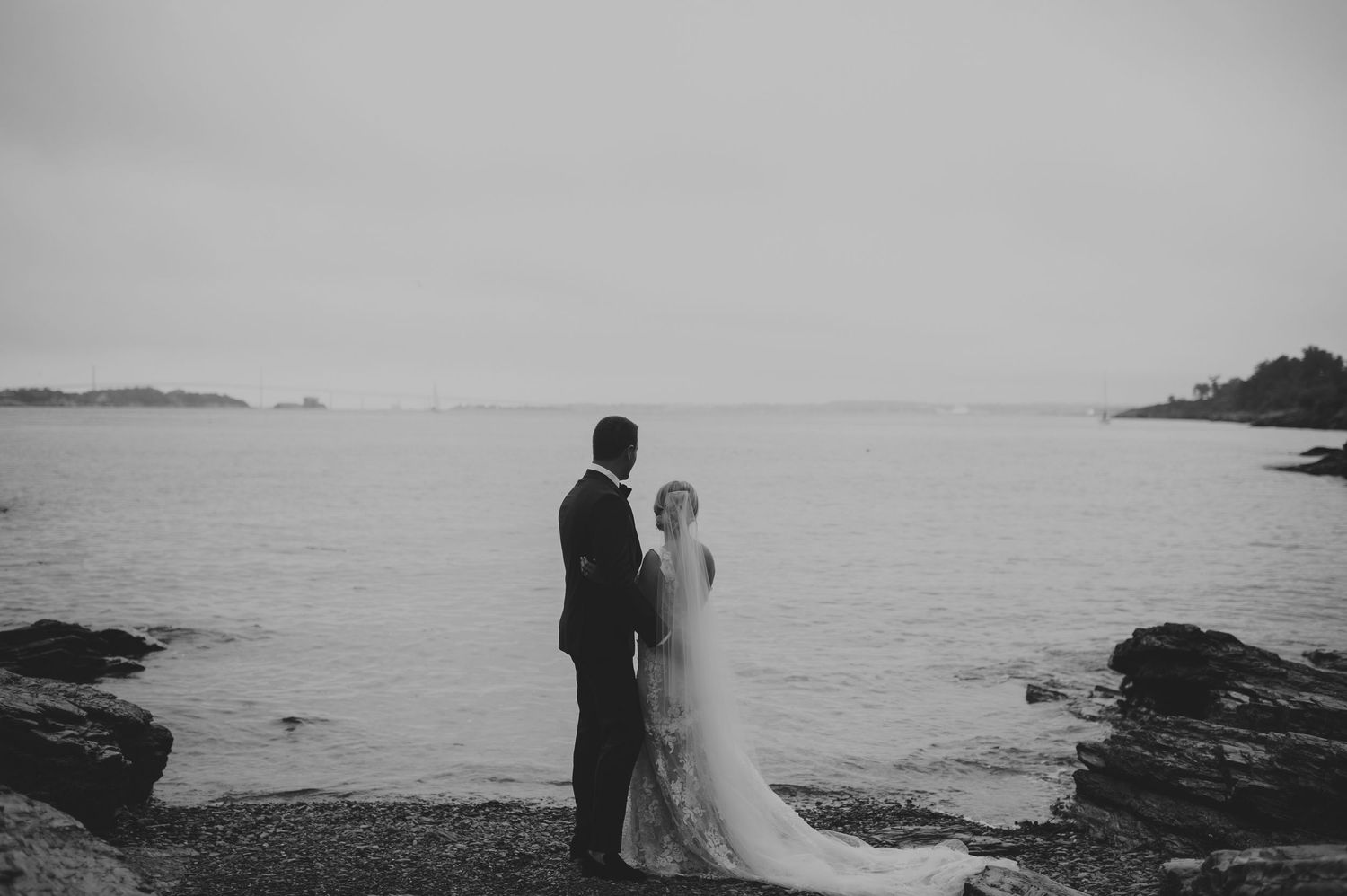 Black and white photo of bride and groom standing together on rocky shore gazing at calm ocean waters.