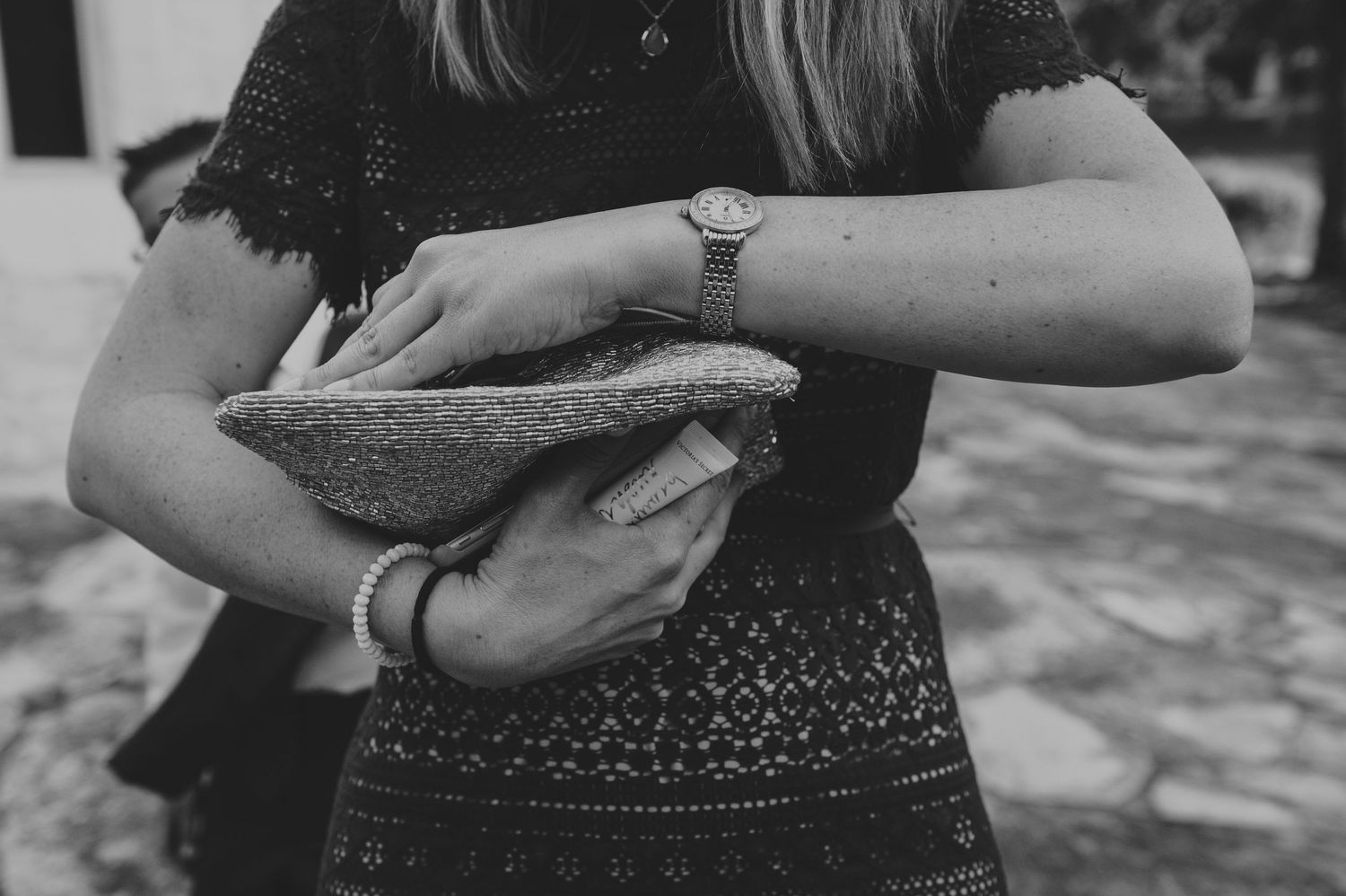 Woman in black lace dress holds a clutch purse while wearing a gold watch.